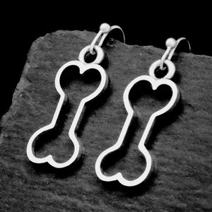 May include: A pair of silver-colored earrings. Each earring features a heart-shaped top and a bone-shaped design. The earrings are displayed on a dark, textured surface, creating a contrast that highlights their shape and shine.
