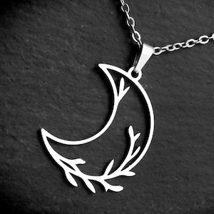 May include: A silver-colored necklace featuring a crescent moon pendant with a delicate leaf design. The pendant hangs from a fine chain, creating a minimalist and elegant aesthetic. The necklace is set against a dark background.