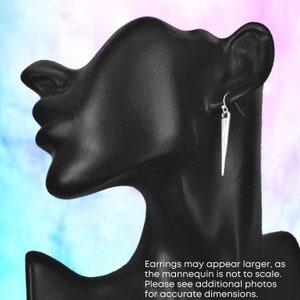 May include: Silver-coloured, pointed dangle earrings displayed on a black mannequin head. The earrings are long and slender, with a hook closure. The background is a gradient of pink, blue, and purple. Text on the image states: "Earrings may appear larger, as the mannequin is not to scale."