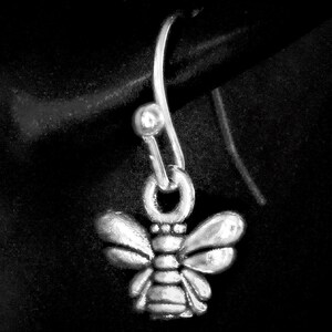May include: A silver-toned bee-shaped earring with a curved hook. The bee has detailed wings and body segments, suspended against a dark background. The earring is a small, delicate piece of jewelry.