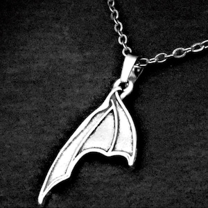 May include: A silver-toned metal pendant shaped like a single bat wing, suspended from a matching silver-toned link chain. The pendant features detailed structural lines and a textured surface, showcasing a pointed tip and a curved wing edge.