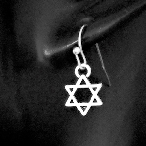 May include: A silver-toned earring featuring a six-pointed Star of David charm suspended from a simple curved hook wire. The metallic star has a clean, open-frame design with sharp geometric points, creating a balanced and symmetrical appearance.