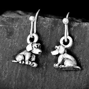 May include: A pair of silver-toned drop earrings featuring small, three-dimensional dog charms. Each charm depicts a seated canine figure with detailed features, suspended from a simple silver-toned hook wire mechanism for pierced ears.