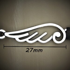 May include: A silver-colored wing charm, 27mm in length, is the focal point of this bracelet. The charm is connected to a black braided cord. The image is a close-up, highlighting the details of the wing design and the cord's texture.