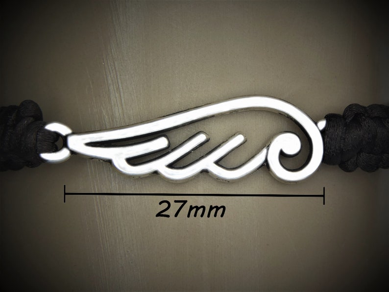 May include: A silver-colored wing charm, 27mm in length, is the focal point of this bracelet. The charm is connected to a black braided cord. The image is a close-up, highlighting the details of the wing design and the cord's texture.