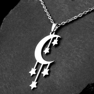 May include: Silver-toned necklace featuring a crescent moon pendant with dangling stars. The celestial-themed jewelry is displayed against a dark, textured background, highlighting the intricate design and metallic sheen.