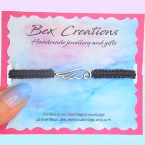 May include: A black macrame bracelet with a silver-colored wing charm. The bracelet is displayed on a card with the text "Bex Creations Handmade jewellery and gifts". The card has a pink border and a blue and white watercolor background.