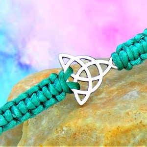 May include: A teal braided bracelet with a silver Celtic knot charm. The bracelet is displayed on a textured, tan surface with a blurred, colourful background. The Celtic knot is a symbol of interconnectedness.