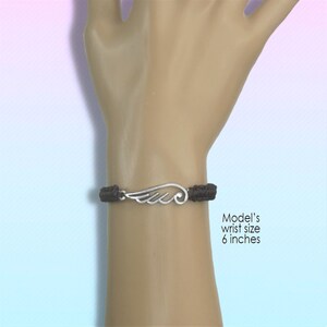 May include: A dark brown braided bracelet with a silver-colored wing charm. The bracelet is shown on a wrist with a 6-inch circumference. The wing charm has detailed feather designs.