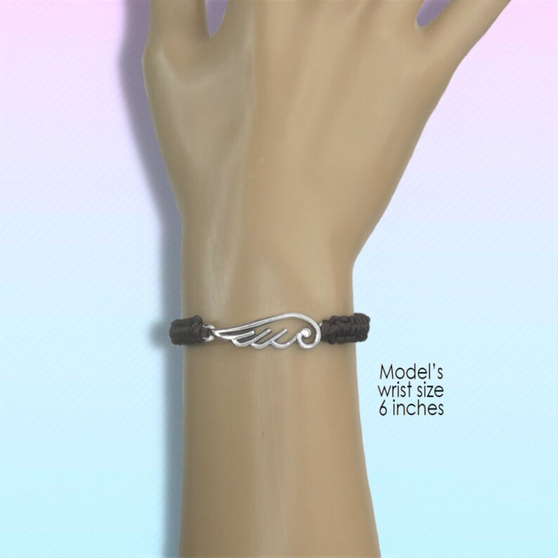 May include: A dark brown braided bracelet with a silver-colored wing charm. The bracelet is shown on a wrist with a 6-inch circumference. The wing charm has detailed feather designs.