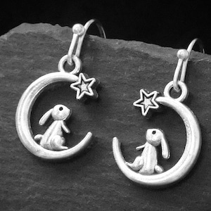 May include: Silver-toned earrings featuring a crescent moon design with a small rabbit figure seated inside. A star charm hangs above the moon. The earrings have a hook closure and are displayed against a dark background.