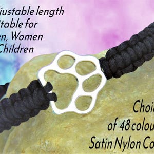 May include: A black braided satin nylon cord bracelet featuring a silver-toned metal paw print charm. The text on the image reads: Adjustable length Suitable for Men, Women & Children and Choice of 48 colours Satin Nylon Cord.
