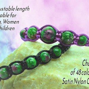 May include: Two adjustable bracelets featuring round green and magenta speckled beads woven into satin nylon cord. One bracelet uses purple cord, the other black. Text reads: Adjustable length Suitable for Men, Women & Children and Choice of 48 colours Satin Nylon Cord.
