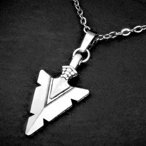 May include: A silver-colored arrowhead pendant necklace. The pendant is detailed with a textured design and is suspended from a delicate chain. The necklace is displayed against a dark background.