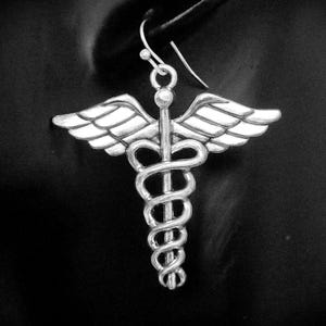 May include: A silver-colored earring featuring the Caduceus symbol, a staff with two snakes and wings. The earring is suspended from a hook and is set against a dark background. The design is detailed and three-dimensional.