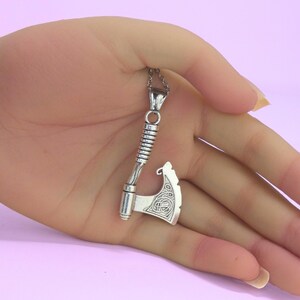 May include: A silver-toned metal pendant shaped like a battle axe, featuring a textured handle and an intricate knotwork pattern on the blade. The pendant is attached to a thin metal chain, showcasing a detailed design with a polished finish.