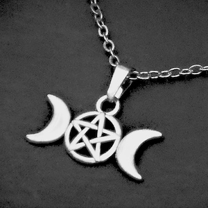 May include: Silver-toned pendant necklace featuring a triple moon design with a central pentagram. The pendant is suspended from a delicate chain. The necklace is displayed against a dark background.