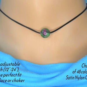May include: A black satin nylon cord necklace featuring a green and red gemstone bead. The necklace is fully adjustable, with a length of 30.5 to 61 cm. The image also shows text that says "Choice of 48 colours".