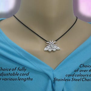 May include: A silver-toned moth pendant suspended on a black cord. The moth features intricate wing patterns and a crescent moon symbol above its head. Text on the image reads: Choice of fully adjustable cord or various lengths and Choice of over 48 cord colours or Stainless Steel Chain.