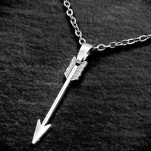May include: A silver arrow pendant necklace. The arrow pendant is detailed with a pointed arrowhead and fletching. The necklace chain is a delicate, linked design. The jewelry is displayed against a dark, textured background.