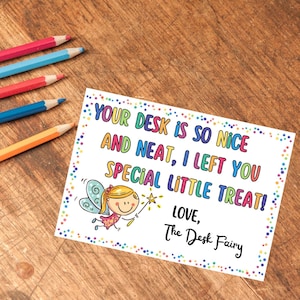 May include: A white card with colorful text and a cartoon fairy illustration. The text reads "YOUR DESK IS SO NICE AND NEAT, I LEFT YOU SPECIAL LITTLE TREAT! LOVE, The Desk Fairy." Colored pencils are in the background.