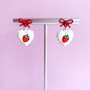 May include: A pair of red bow earrings with white heart-shaped charms featuring a red strawberry design.