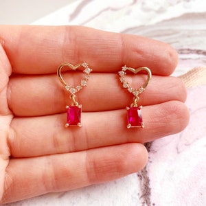 May include: Gold heart shaped earrings with a dangling red square gem. The heart is outlined with small clear stones.