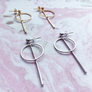 May include: A pair of gold and silver earrings with a circle and a long bar design. The earrings are on a white marble surface.