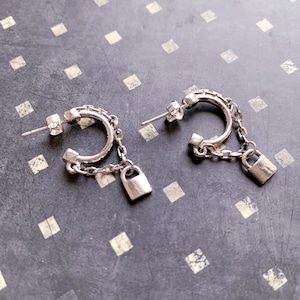 May include: A pair of silver hoop earrings with a dangling padlock charm and a small silver chain. The earrings are on a gray background.