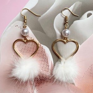 May include: A pair of gold heart-shaped earrings with white faux fur pom-poms and white pearl accents.