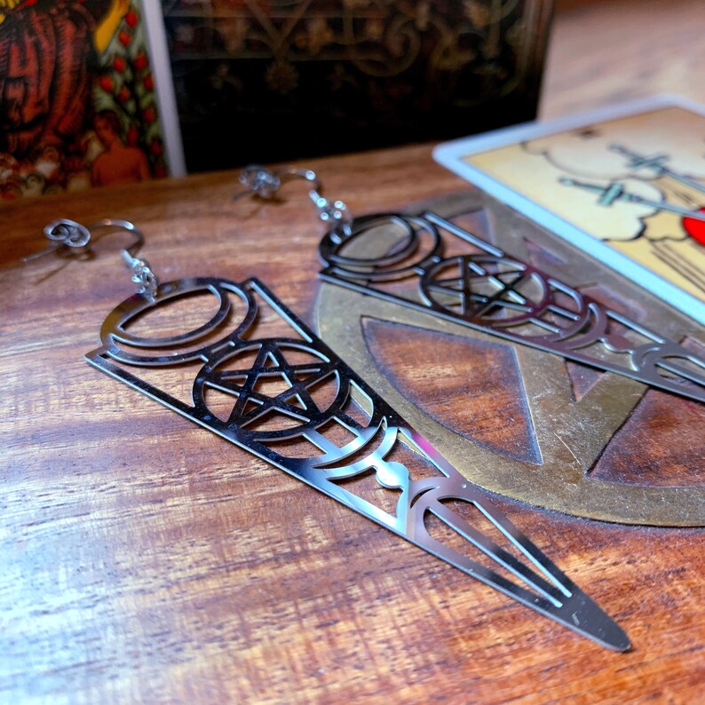Puede incluir: Un par de pendientes de plata con un dise&ntilde;o de pentagrama y fases lunares. Los pendientes est&aacute;n hechos de metal y tienen una forma puntiaguda.
