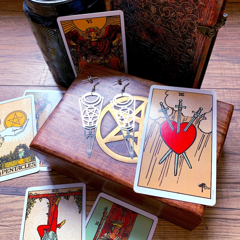 Puede incluir: Dos pendientes de plata con un dise&ntilde;o de pentagrama, expuestos en una caja de madera con cartas de tarot. Las cartas muestran el 'Siete de Pent&aacute;culos' y el 'Tres de Espadas'.