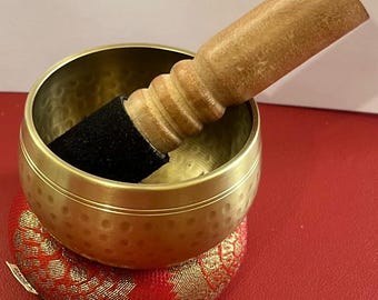 May include: A gold singing bowl with a wooden striker and a red cushion. The bowl has a hammered texture and a smooth rim.