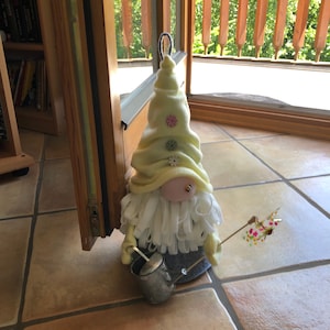 Könnte beinhalten: Ein gelber Filz-Zwerg mit weißem Bart und einem grauen Filzhut. Der Zwerg hält eine kleine Gießkanne aus Metall und einen Stock mit einer bunten Blumenverzierung.