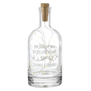 Może przedstawiać: Przezroczysta szklana butelka z korkowym korkiem i sznurem światełek w środku. Na butelce wygrawerowano napis "Der schönste Weg ist der gemeinsame" wraz z sercem i papierowym samolotem. Na butelce wygrawerowano również imiona Stefanie & Lennart.