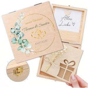 Op de afbeelding: Een lichtgekleurde houten bewaardoos met een gouden cirkelvormig frame, eucalyptusbladeren, twee gouden harten en de tekst "unsere Hochzeit Thomas & Sandra 10.07.2026". De binnenkant bevat een kaart met "Alles Liebe" en een houten cadeau-uitsparing.