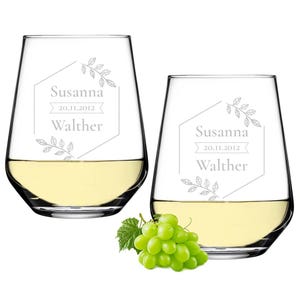May include: Two clear glass wine glasses with a white wine inside. Each glass has a personalized inscription with the name "Susanna Walther" and the date "20.11.2012" in a geometric frame with a leafy design.