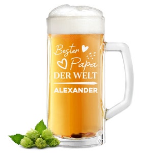 Könnte beinhalten: Ein klares Glasbierkrug mit einer weißen Aufschrift, die "Besten Papa der Welt Alexander" lautet. Der Krug ist mit goldenem Bier gefüllt und hat einen weißen Schaumkopf. Im Vordergrund befinden sich grüne Hopfendolden.
