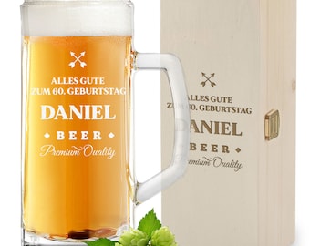 Op de afbeelding: Een heldere glazen bierpul gevuld met goudkleurig bier en wit schuim, voorzien van de tekst ALLES GUTE ZUM 60. GEBURTSTAG DANIEL BEER Premium Quality. Daarnaast staat een lichthouten kist met dezelfde tekst en groene hopbellen.