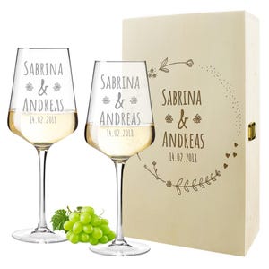 Puede incluir: Dos copas de vino de vidrio transparente con el texto "Sabrina & Andreas 14.02.2018" grabado en ellas. Las copas están en una caja de madera con el mismo texto y un diseño floral.