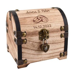 Op de afbeelding: Houten schatkist met metalen accenten en een hartvormig slot. De kist is gegraveerd met de namen "Anna & Peter", trouwringen en de datum "14.12.2022". De kist heeft zwarte leren banden en messingkleurige hardware.
