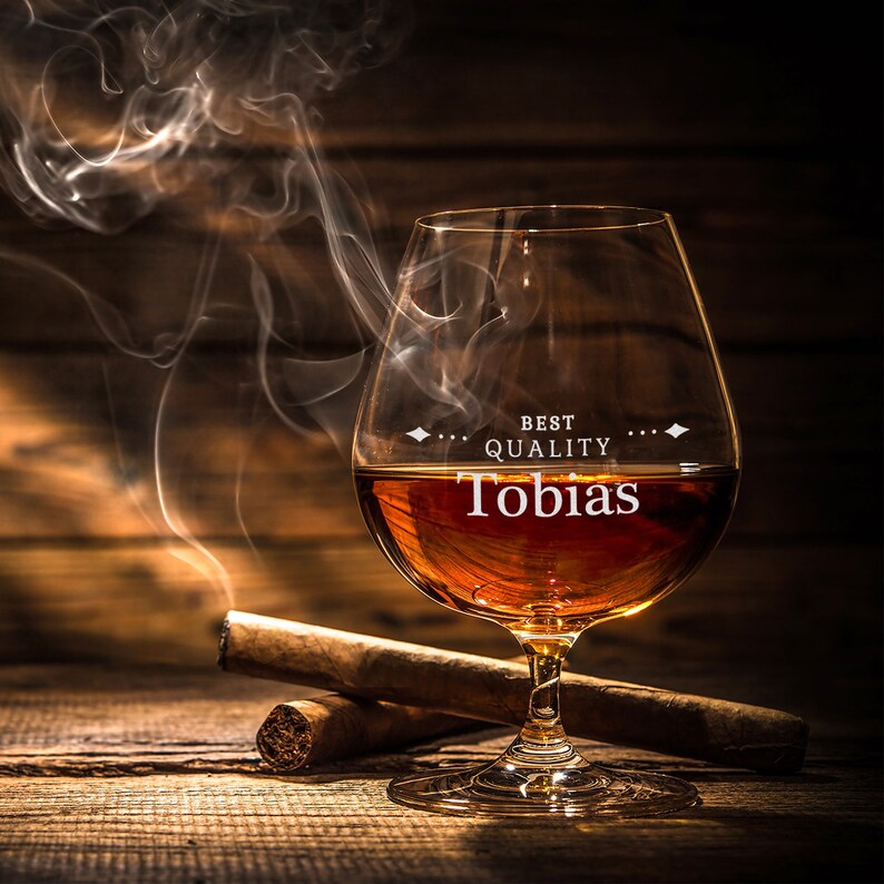May include: A brandy glass with amber liquid, the name "Tobias" printed on the glass, and the words "BEST QUALITY" above it. Two cigars lie on a wooden surface, with smoke rising from the glass. The image has a warm, inviting tone.