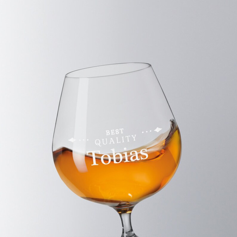 May include: A clear brandy glass with the text "BEST QUALITY Tobias" etched on the glass. The glass is filled with amber liquid, with a splash visible. The glass has a rounded bowl and a short stem.
