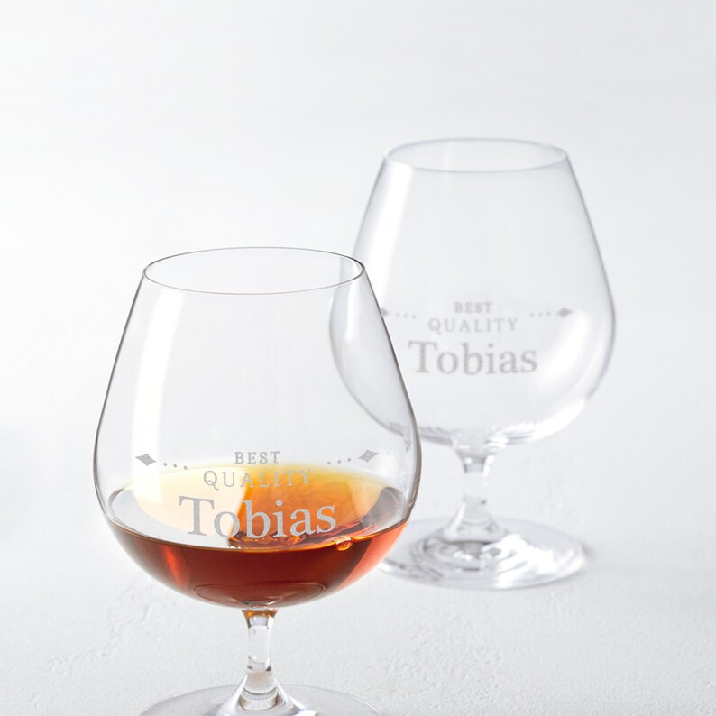 May include: Two clear brandy glasses with the text "BEST QUALITY Tobias" etched on the glass. One glass is partially filled with a brown liquid. The glasses have a rounded bowl and a stemmed base.
