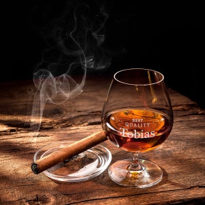May include: A brandy glass with amber liquid, engraved with "Best Quality Tobias", is on a wooden surface beside a lit cigar in a clear ashtray. Smoke curls upwards from the cigar, creating a dramatic effect.