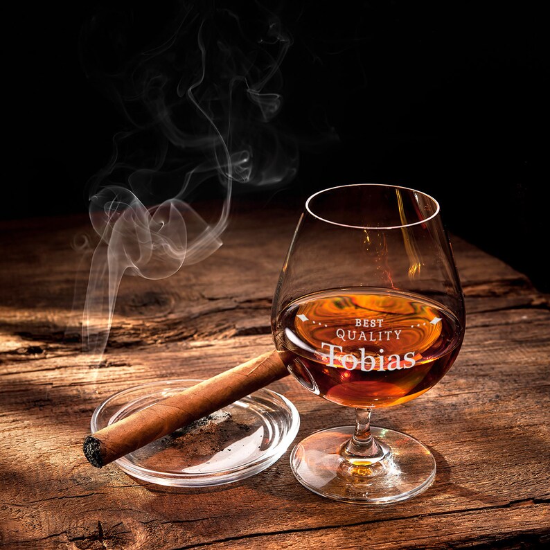 May include: A brandy glass with amber liquid, engraved with "Best Quality Tobias", is on a wooden surface beside a lit cigar in a clear ashtray. Smoke curls upwards from the cigar, creating a dramatic effect.