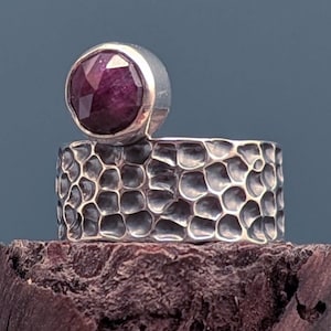 Puede incluir: Un anillo de plata con una gran gema facetada de color rojo intenso. La banda del anillo tiene un patrón texturizado en forma de panal. La gema está engastada en un engaste de plata redondeado y elevado.