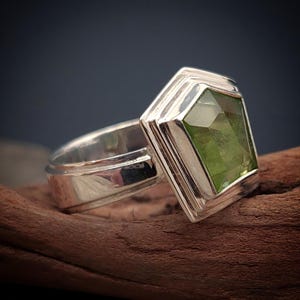 Könnte beinhalten: Ein silberner Ring mit einem grünen Edelstein, der in einem geometrischen Design gefasst ist.