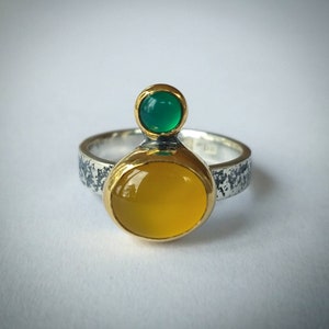 May include: A silver ring with a textured band, featuring a gold-plated setting holding a large, oval yellow gemstone and a smaller, round green gemstone. The ring is set against a white background.