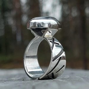 May include: A polished silver ring featuring a wide, thick band with deep, diagonal etched grooves along the exterior. The ring is topped with a large, smooth, dark, rounded stone setting that sits prominently above the band.